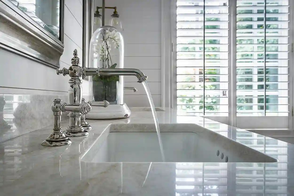 Luxurious kitchen sink with classic silver faucet, marble countertop, and a view to greenery through white shutters.