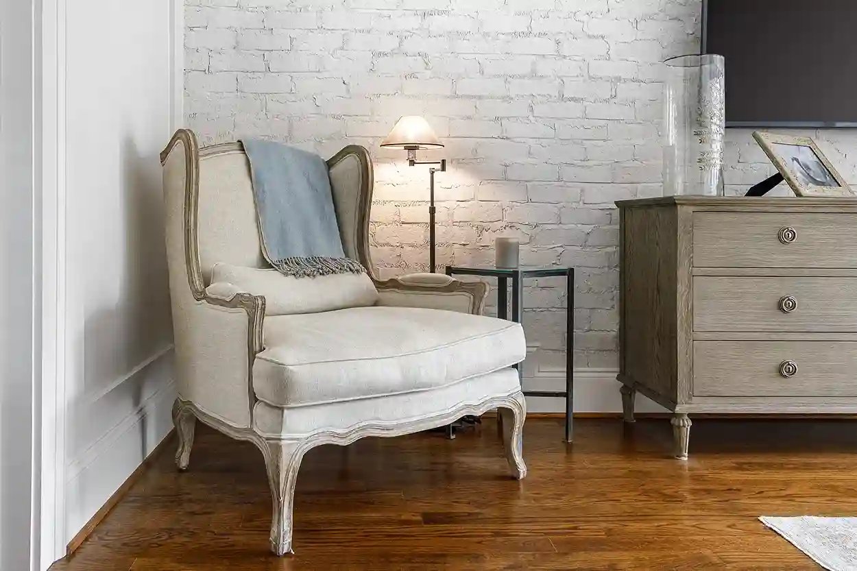 Vintage-inspired armchair with throw blanket beside a distressed dresser and floor lamp against a white brick wall