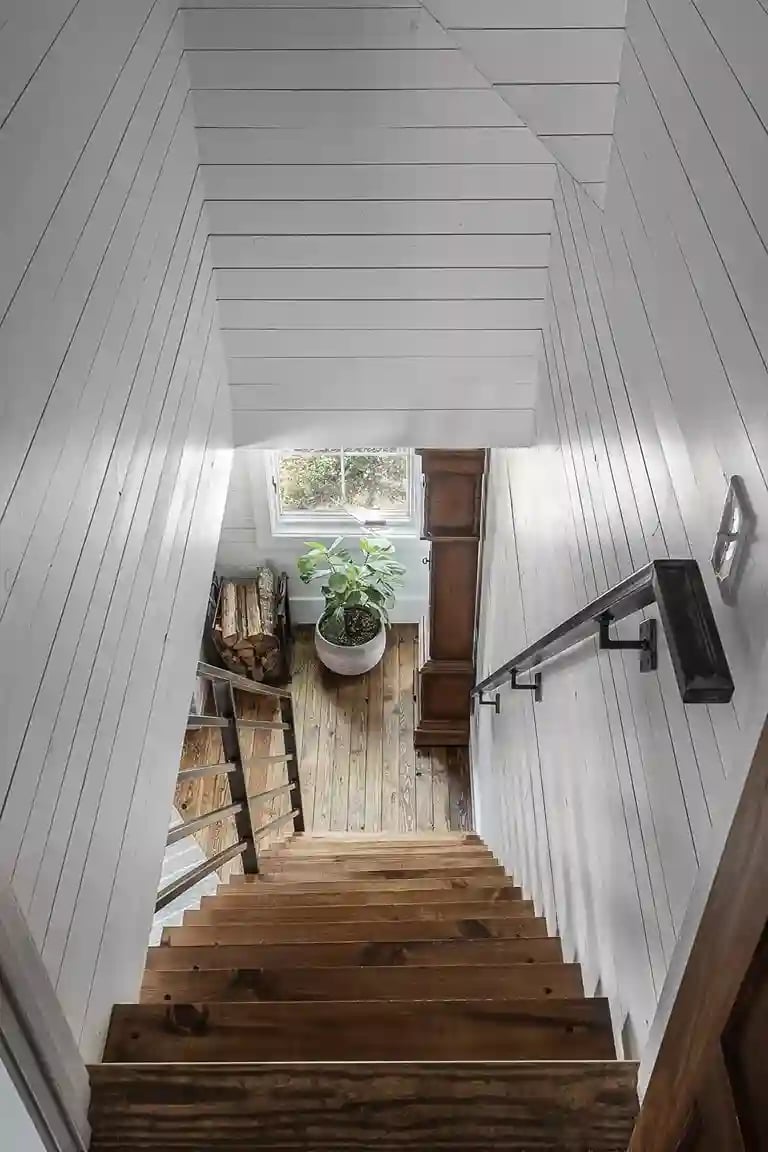 Downward view of a rustic wooden staircase with white shiplap walls and a potted plant at the base.