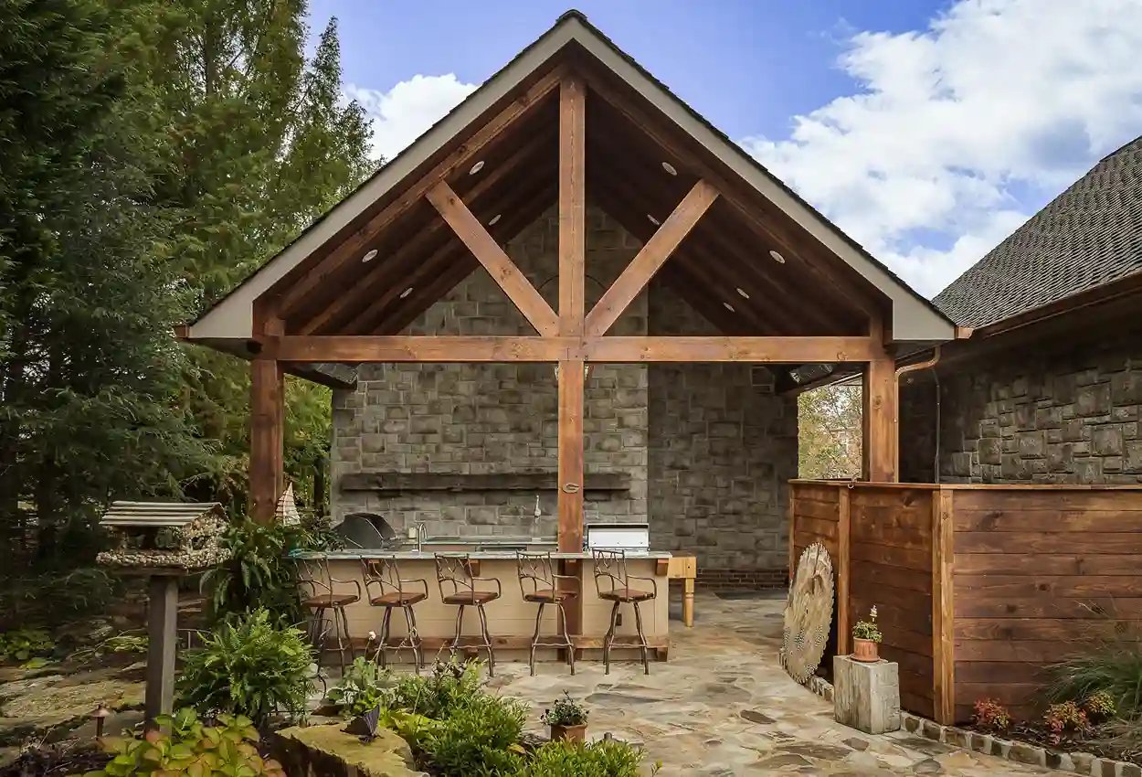  Outdoor kitchen pavilion with stone walls, wooden beams, and bar seating in a garden setting.