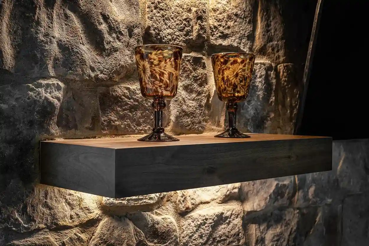 Elegant amber glass goblets on a floating wooden shelf against a textured stone wall.