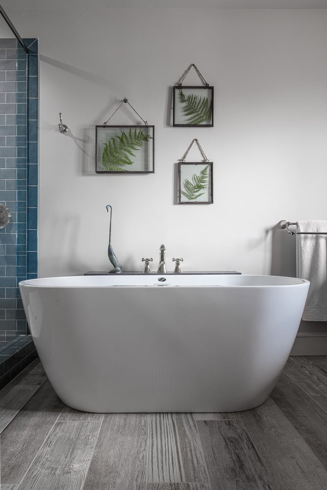 Elegant freestanding bathtub with wall-mounted faucet, framed fern artwork, and blue tile accent.