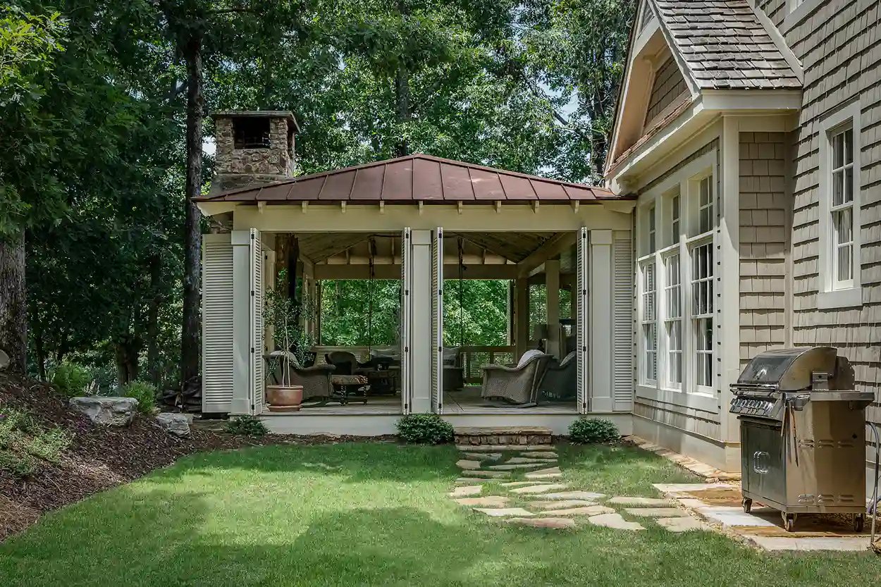 Covered outdoor patio area with comfortable seating and grill, adjacent to a shingled home surrounded by lush greenery.