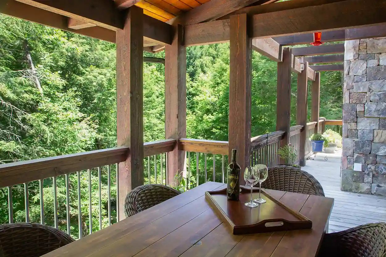 Wooden patio dining area with a rustic table set, wine bottle, and glasses overlooking dense greenery.