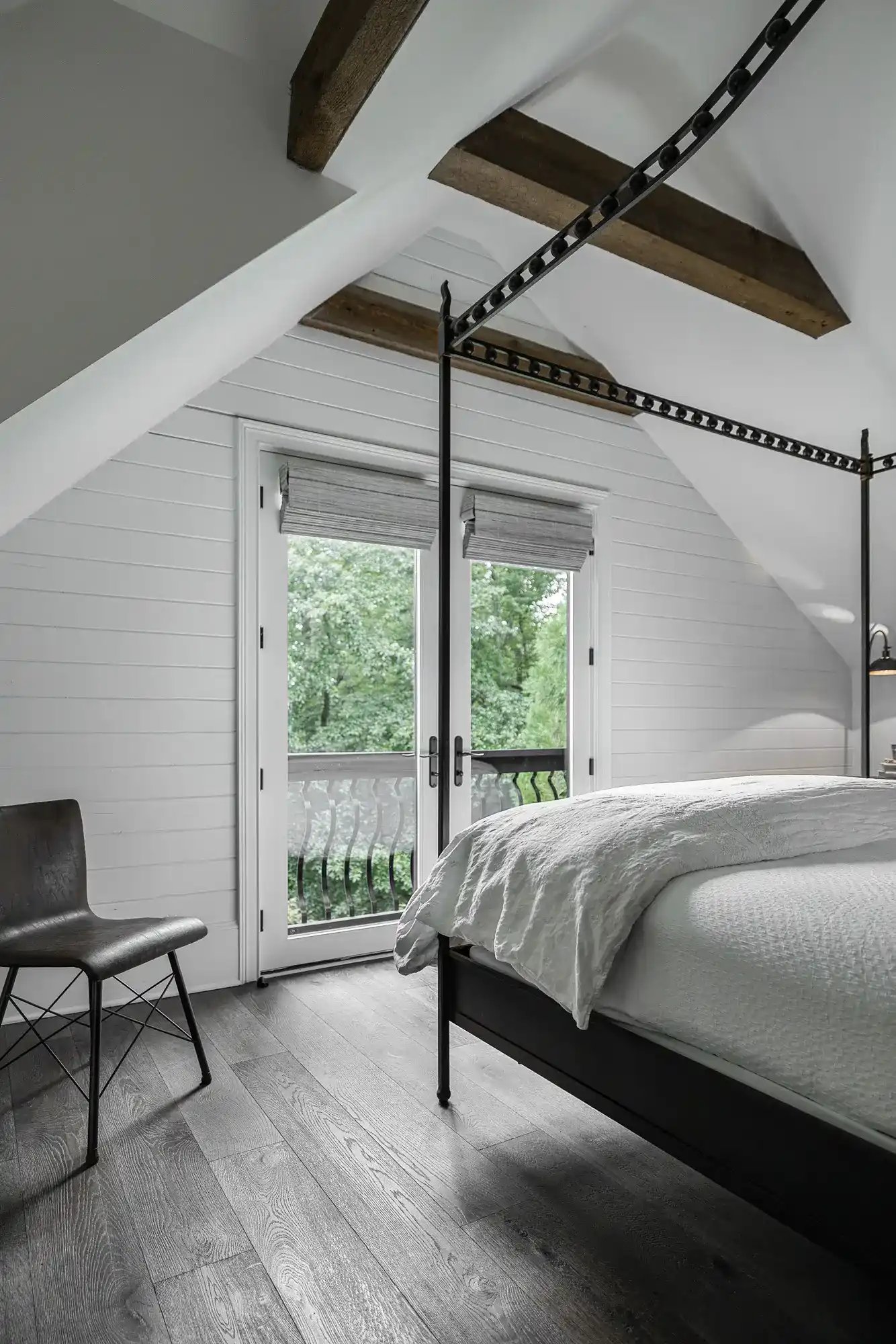 Minimalist bedroom with exposed beams, shiplap walls, and French doors opening to a lush view.
