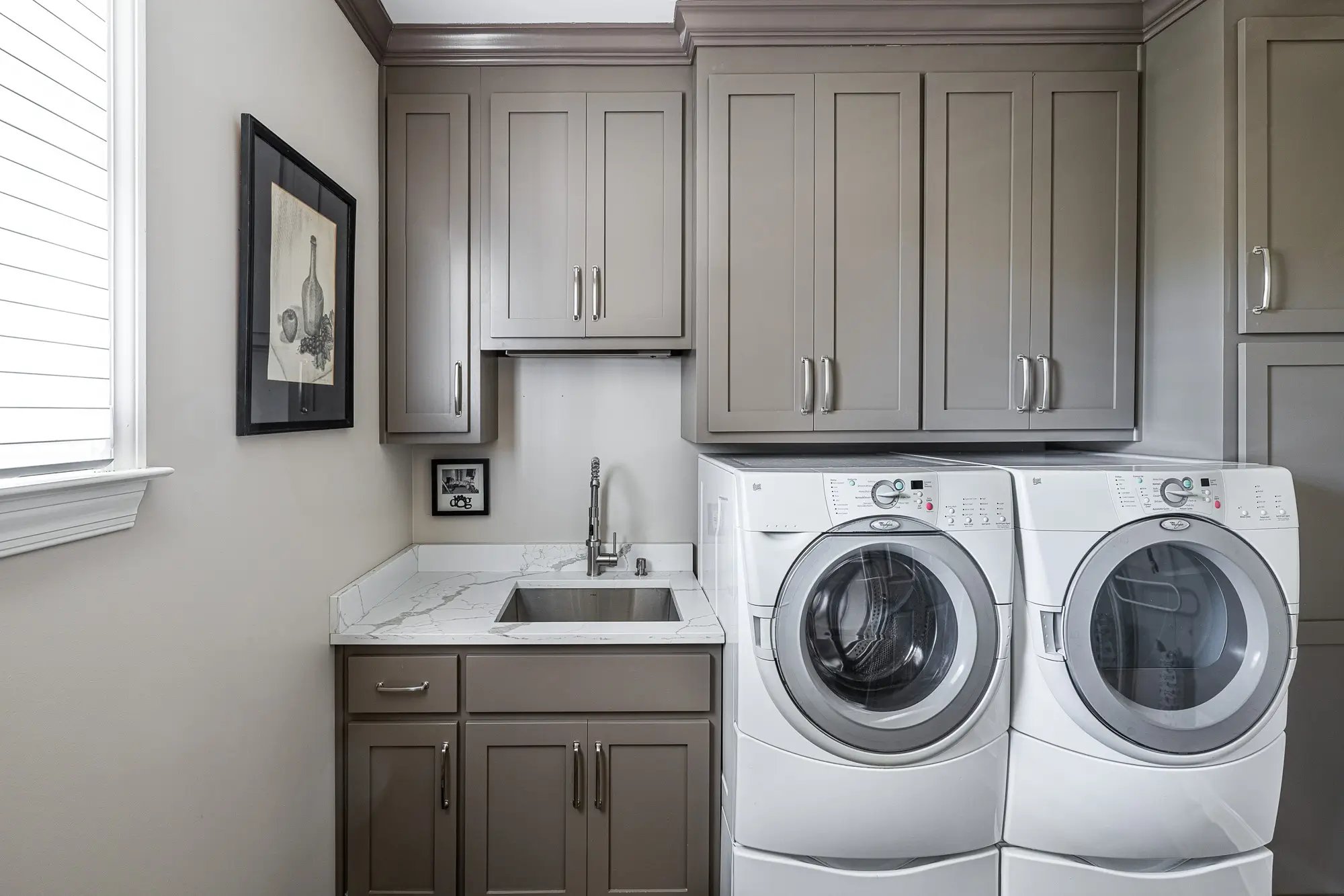 Modern laundry room with taupe cabinets, marble countertops, and high-efficiency front-load washer and dryer.