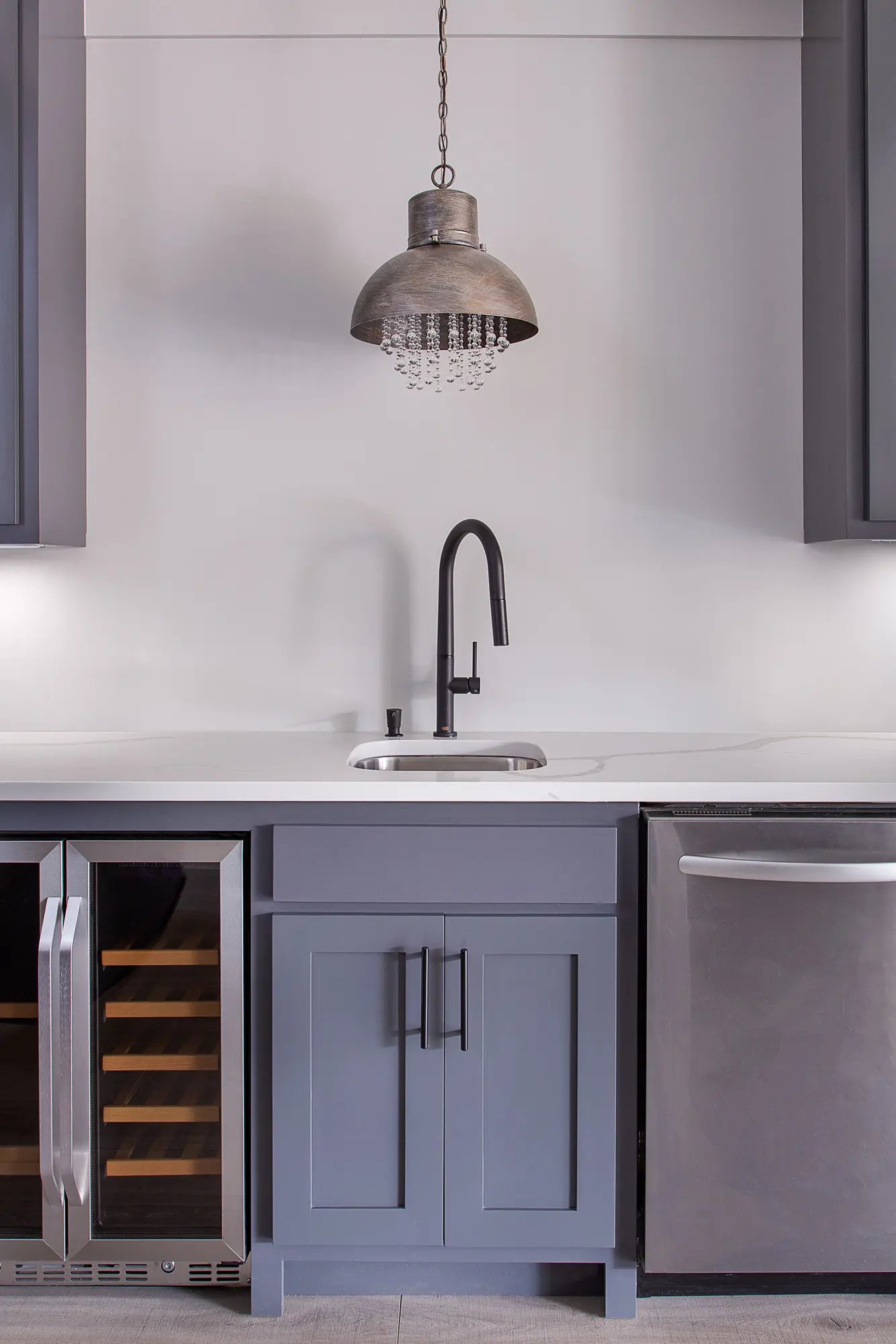 Elegant kitchen wet bar with gray cabinetry, white countertop, undermount sink, black faucet, and industrial pendant light.