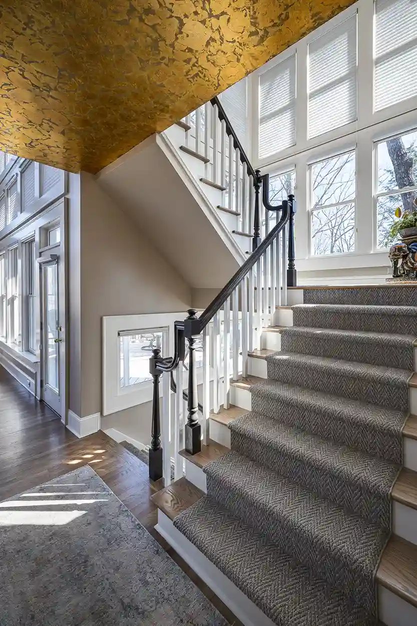 Luxurious staircase with textured carpet, gold leaf ceiling, and abundant natural light from tall windows.