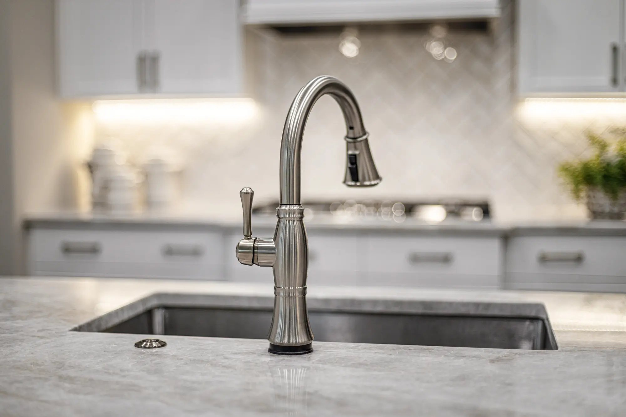 Elegant kitchen faucet over a granite sink with a blurred herringbone tile backsplash and white cabinetry.