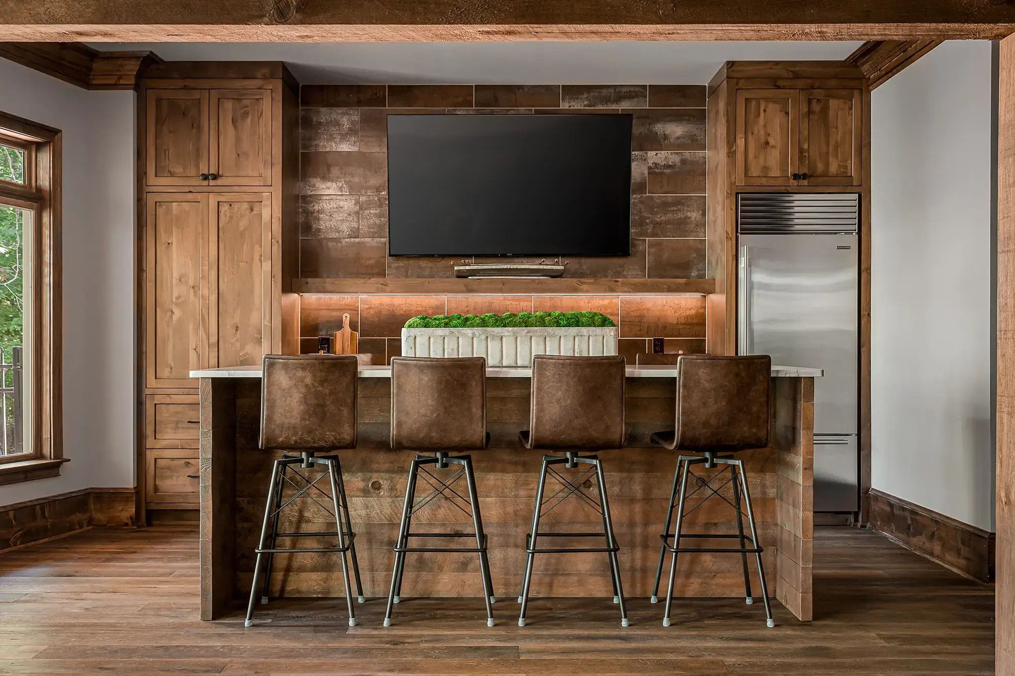 Rustic home bar with leather stools, wooden cabinetry, large TV, and refrigerator in a cozy setting.