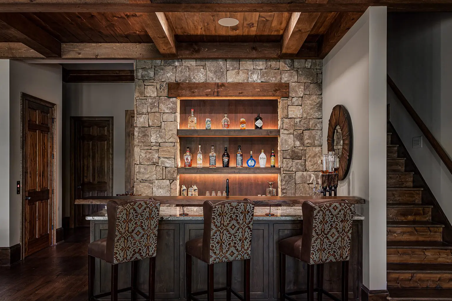 Home bar with stone accent wall, wooden shelves for liquor display, and patterned bar stools.