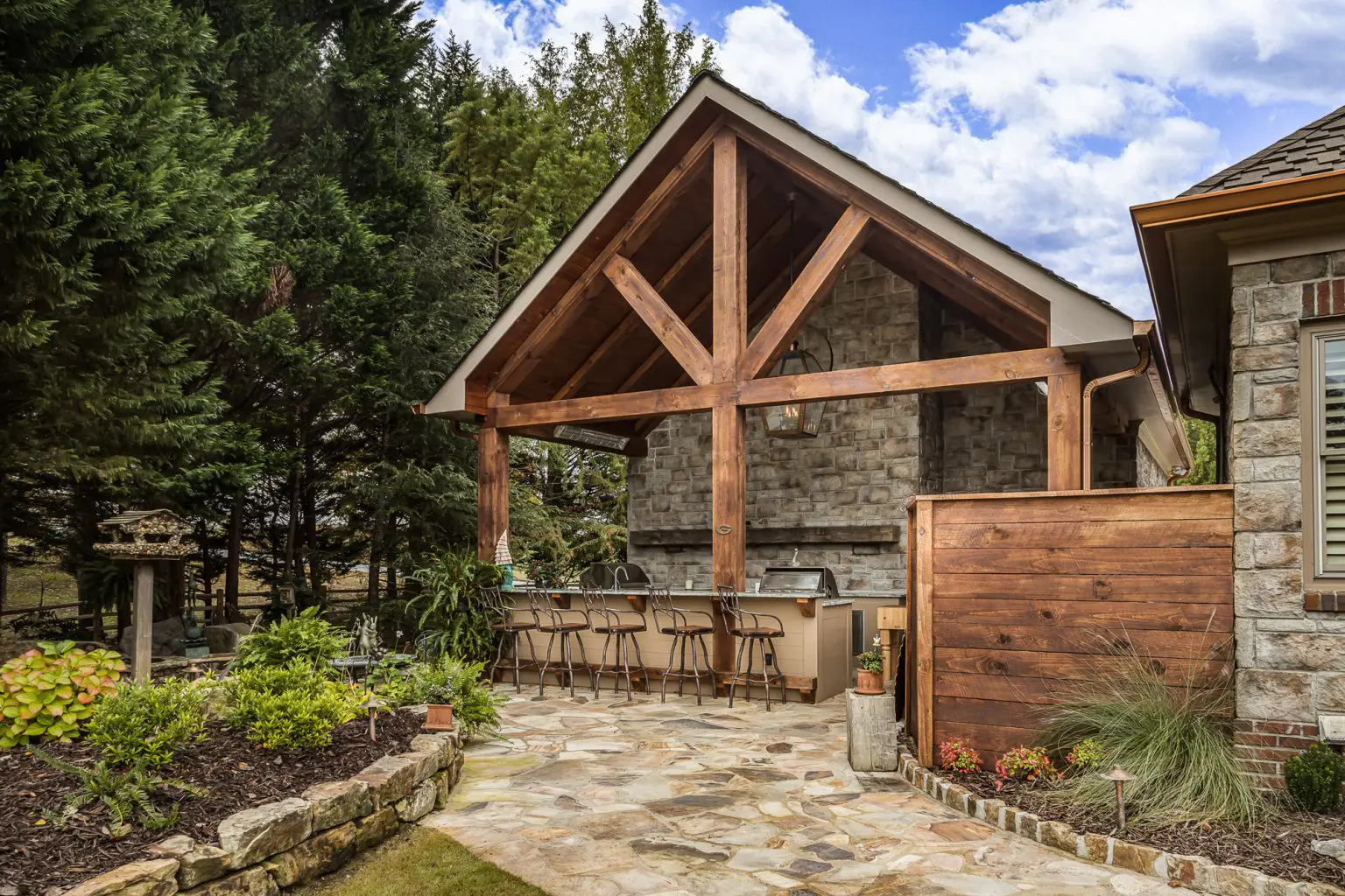 Outdoor kitchen pavilion with stone walls, wooden beams, and bar seating in a garden setting.